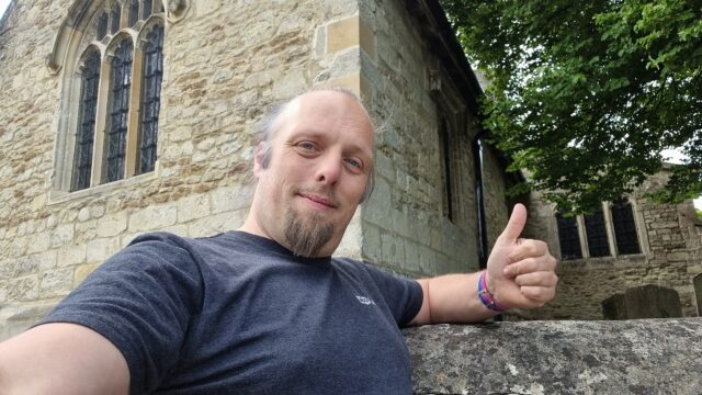 Outside a Norman-style church, Dan throws a thumbs-up sign.
