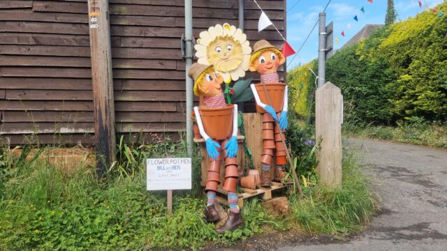 Human-sized scarecrows of classic childrens' TV characters Bill and Ben the Flower Pot Men and their friend Little Weed, constructed mostly out of plant pots, standing at the corner of a road by a wood-panelled outbuilding.