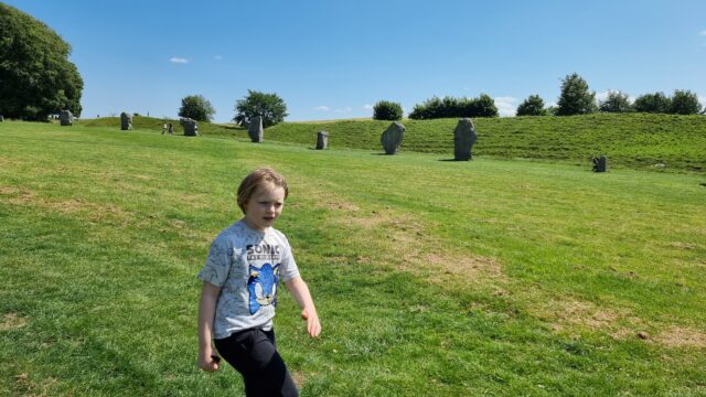 On verdant grass and under clear skies, a boy in a white-and-blue 'Sonic the Hedgehog' t-shirt strolls in front of a line of about half a dozen enormous standing stones.