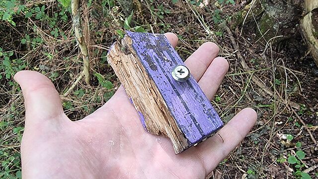 A hand holds a palm-sized fragment of wood, painted purple as if once part of a fairy door.