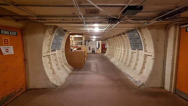 A circular tunnel with a flat floor and low ceiling, wrapped in white-painted iron and concrete, with exposed wiring and WWII-era signage, leads deeper into a subterranean bunker past a small guard post in which a figure can barely be seen, reading a book.