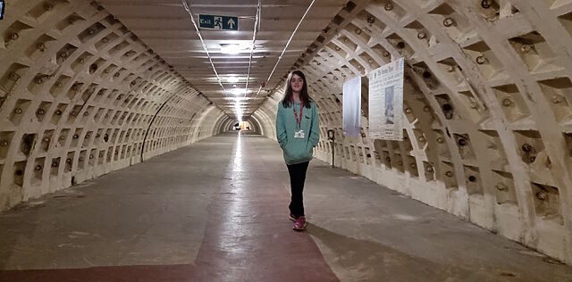 In an extremely long semicircular concrete tunnel, a tween girl wearing a green hoodie stands near signage made to look like 1940s newspapers.