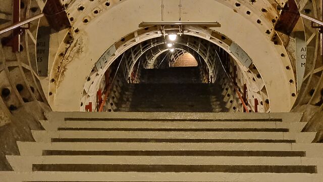 Broad staircase reminiscent of those found around the London Underground, but sparsely lit and decorated. Looking up the stairs, it ends in a brick wall.