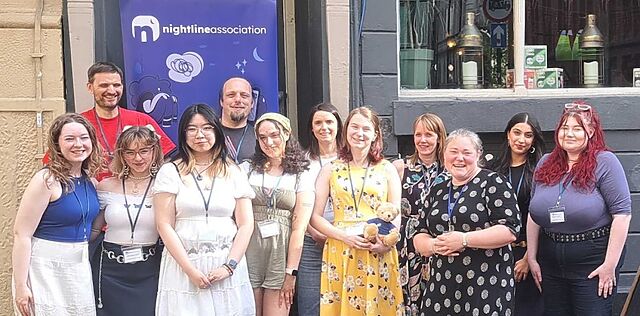 12 Caucasian people of a mix of ages and genders posing as a group in front of a Nightline Association banner. Dan is one of them.