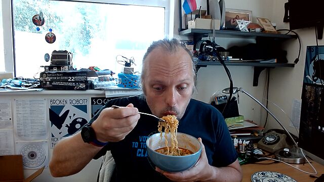 Dan slurps a forkful of noodles lifted from a bowl full of noodles in a deep red sauce, in a cluttered office space.