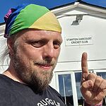 Dan, a white man with a beard, wearing a rainbow bandana, points to a white building behind him.