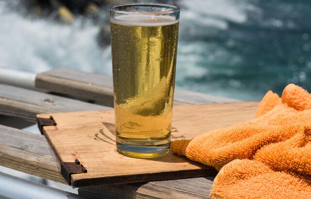 A pint of light-coloured beer on wooden board, with the sea in the background. An orange towel sits alongside.