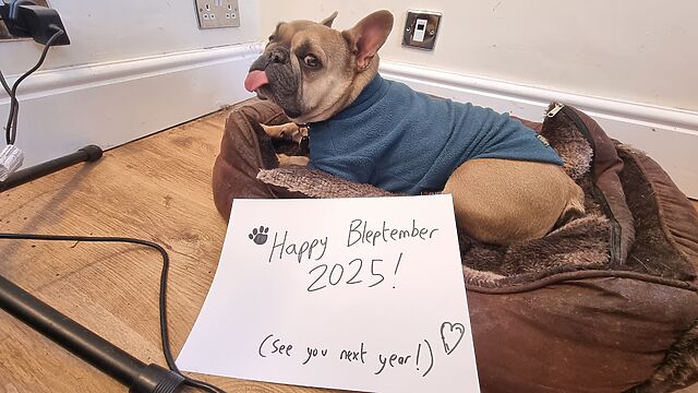 A champagne-coloured French Bulldog in a teal jumper lies in a soft brown dog bed in the corner of an office. She looks over her shoulder at the photographer, her tongue sticking pretty-much entirely out in a long blep. In front of her, a handwritten sign is marked with a pawprint and a heart and the words 'Happy Bleptember 2025! See you next year!'