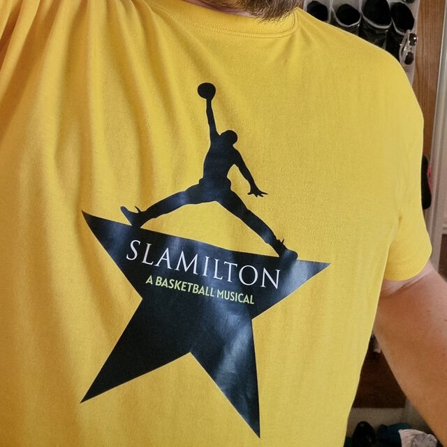 Close-up of a human torso wearing a yellow t-shirt, on which there's a black outline of a five-pointed star with the top point replaced with the silhouette of a person, in the style of the Hamilton logo except that instead of being Alexander Hamilton with his hand held high the silhouette is a basketball player reaching for a leaping dunk. The text on the star reads "Slamilton: A Basketball Musical".