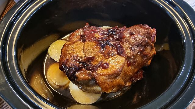 A seared shoulder of pork rubbed with spices sits in a syrupy cider mix atop some sliced white onions in a slow cooker pot.