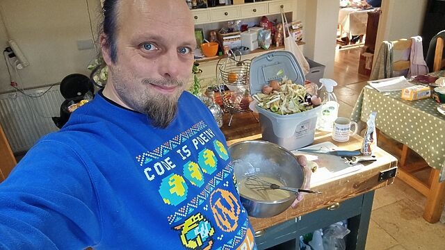 Dan, a white man with a beard and blue hair, wearing a WordPress-themed Christmas jumper, beats a bowl of batter.