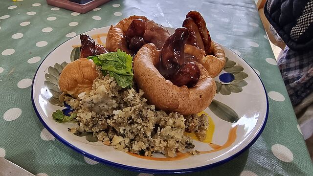 Four Yorkshire puddings on a side plate, each with one or two chipolata sausages sticking out, smothered in gravy, alongside a small quantity of sage & onion stuffing.