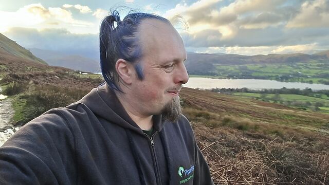 Dan, a white man with blue hair, looks over the valley of the Ullswater.