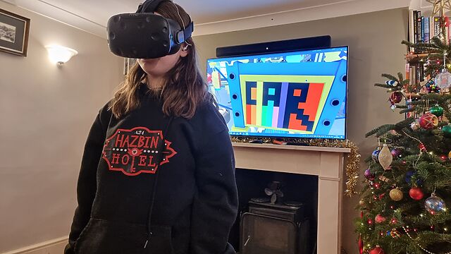 A tweenage girl in a black 'Hazbin Hotel' hoodie wears a VR headset; the screen behind her shows that she's drawn a picture featuring a rainbow background and the word 'CAR', while playing Job Simulator.