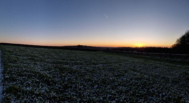 The sun peeks through clouds at the horizon beyond a wide valley full of fields.