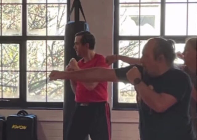 In a gym environment with punchbags visible in the background Dan, a white man wearing a black t-shirt, throws a right jab, alongside others who are doing the same.