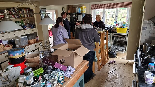 A group of adults stand talking in a disrupted kitchen, with food, furniture, and boxes stacked high.