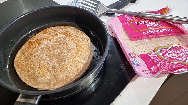 Two layers of chapattis in a frying pan on a stove, alongside their packaging.
