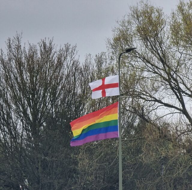 Two flags hang from a tall lamp post. The smaller upper one is a St. George's Cross, the flag of England.the lower, larger (and much nicer) one is the six-colour Pride rainbow.