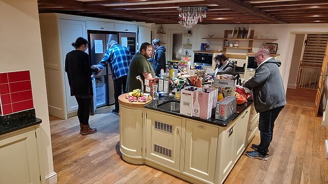 A large group of people stand around a pile of shopping in the centre of a nicely decorated kitchen.