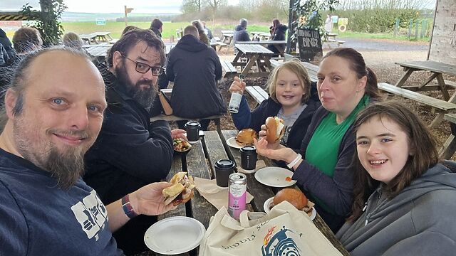 A family sits at a picnic table in a barn, eating sausage sandwiches.