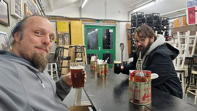 Dan and JTA sit with pints of beer in a cluttered brewery, at a table with large tomato cans repurposed as holders for chilli oil and pizza cutters.