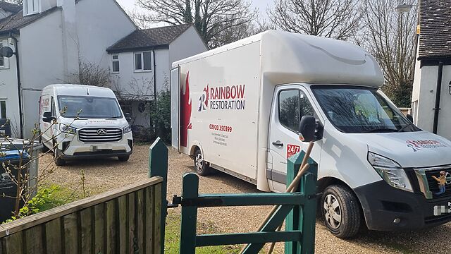 A large van and a small van from Rainbow Restoration sit on a gravel driveway.
