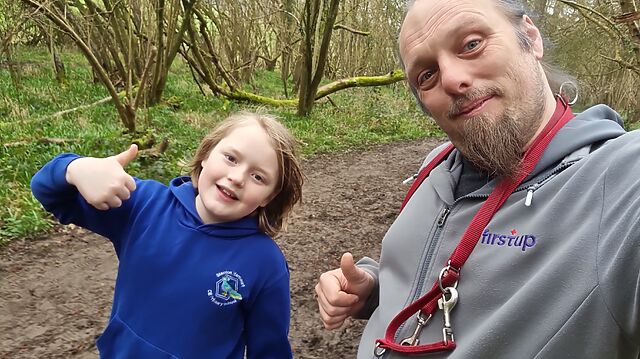 Dan and a boy each throw a thumbs-up to the camera from a forest footpath.