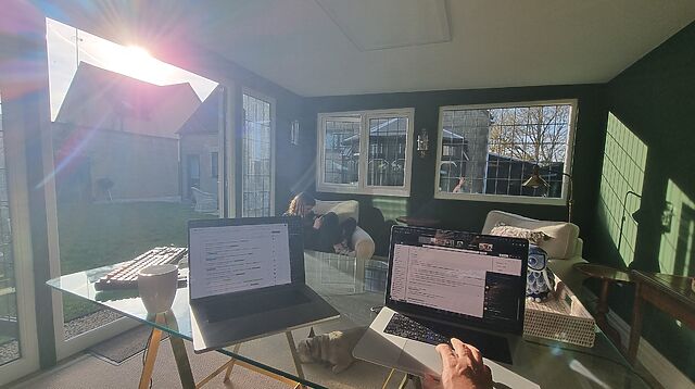 Two laptops on a glass desk in an attractive garden office/summer house, bathed in bright afternoon sunshine.