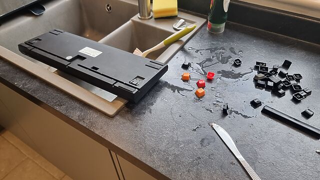 A desktop computer keyboard inverted over a kitchen sink, with many of its keycaps scattered around.