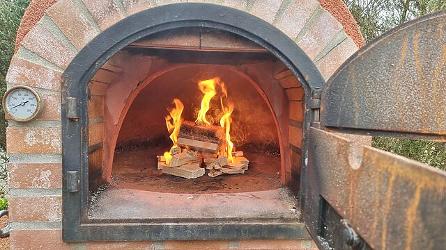 A small wood fire burns inside an outdoor brick pizza oven.