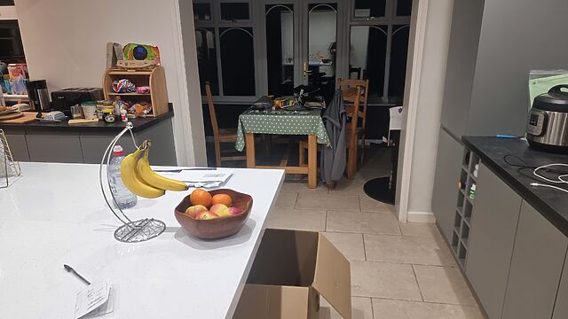 A moderately tidy kitchen with faux-marble countertops, under free on which an open large cardboard box can be seen. In the distance, a conservatory contains a small dining table cluttered with computer equipment.
