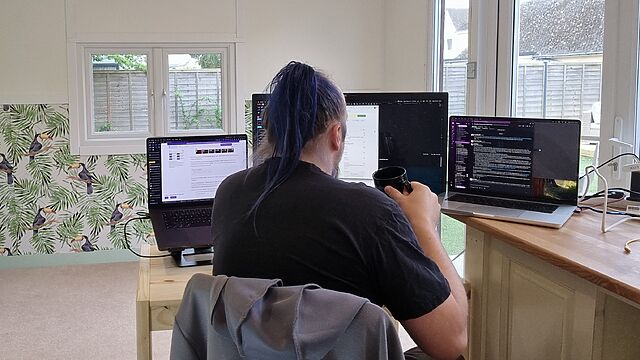 A white man with blue hair tied up in a ponytail sits at a basic pine desk in a garden office, decorated with wallpaper showing toucans. In front of him are two laptops and a large monitor. He holds a black mug in his hand, as if about to drink from it.