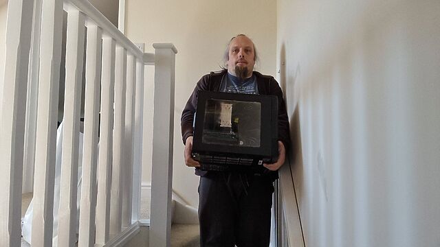 Dan, a white man with a ponytail and a goatee beard, carries a large black cube-shaped computer down a staircase.