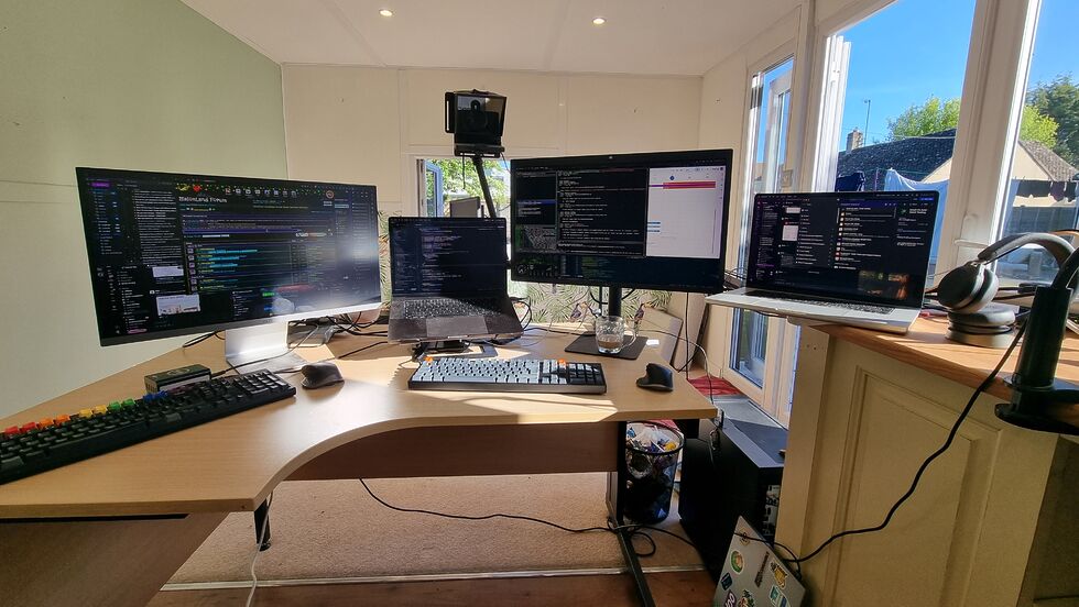 Two laptops sit nestled between two large monitors, with a desktop computer on the floor below and a teleprompter/webcam on an arm above, in a brightly-sunlit garden office.