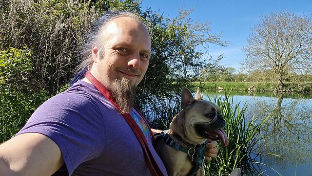 A white man with a blue ponytail and a goatee beard sits with a French bulldog alongside a wide, calm river.