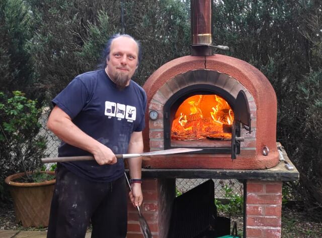 Dan, holding a pair of pizza peels, stands before the roaring fire of the open brick pizza oven, with a pizza barely visible within.