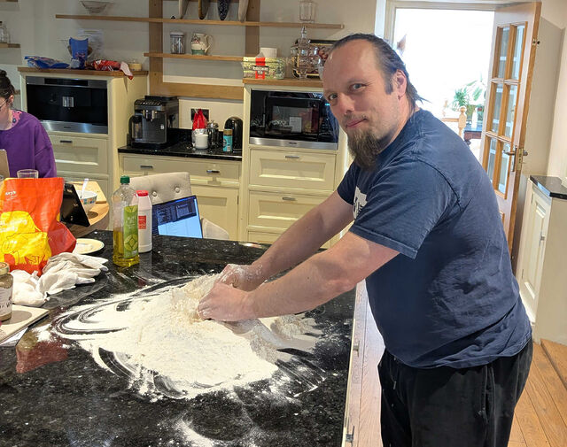 Dan, a white man with a ponytail of blue hair and a goatee beard, uses his hands to gather a huge pile of flour on a marble worksurface in a spacious kitchen.