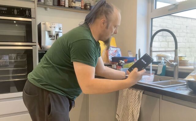 Dan, a white man with a blue ponytail, wearing a green t-shirt, watches a stopwatch timer on his phone while filling a measuring jug from the kitchen sink's tap.