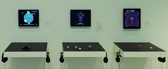 Exhibition space showing wall-mounted cabinet controls for three retro video games, from the header of Mike Cook's original article.
