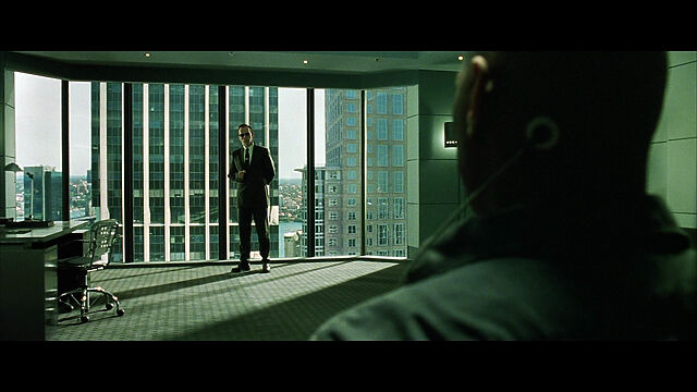 Framegrab from The Matrix. In the foreground is the silhouette of Morpheus, who is about to be interrogated by Agent Smith, a man in a suit at the windowed far end of an office.