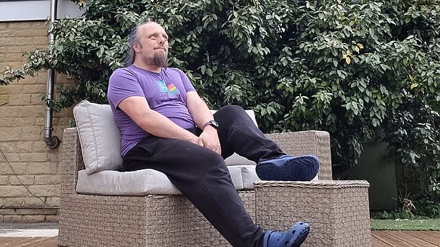Dan, a white man with a blue ponytail and a goatee beard, wears purple t-shirt, tracksuit bottoms, and crocs to sit outdoors on garden furniture in light rain.