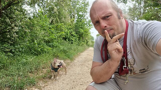 Dan, a white man with a goatee beard and a blue ponytail, looks thoughtful as he crouches on a footpath near a fawn-coloured French Bulldog.