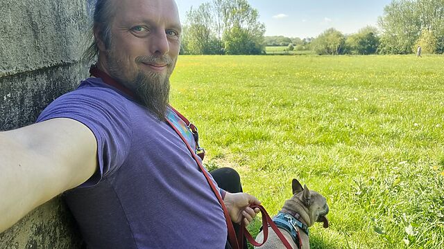 Dan, a white man with a blue ponytail and a goatee, sits in the shadow of an old concrete wall alongside a leashed French Bulldog, with a large sun-dappled meadow visible in the background.