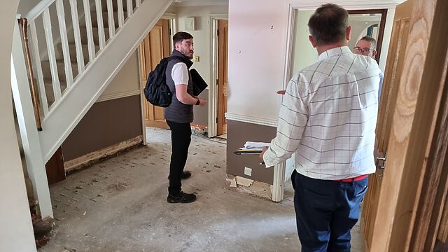 Several men stand on the bare concrete floor of a residential hallway with no furniture or skirting boards.