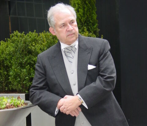 Carl Person, an older white man with receding grey-white hair, wearing a smart three-piece suit, leans against a planter in an marbled ornamental garden.