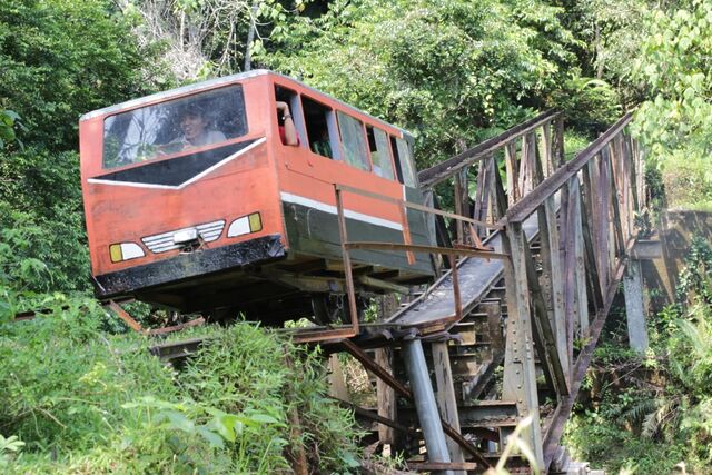 A tiny enclosed passenger rail vehicle crosses an old narrow iron bridge in a jungle.