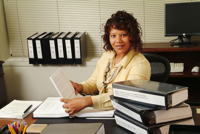 A brown-skinned woman sits at a desk surrounded by binders of paperwork.