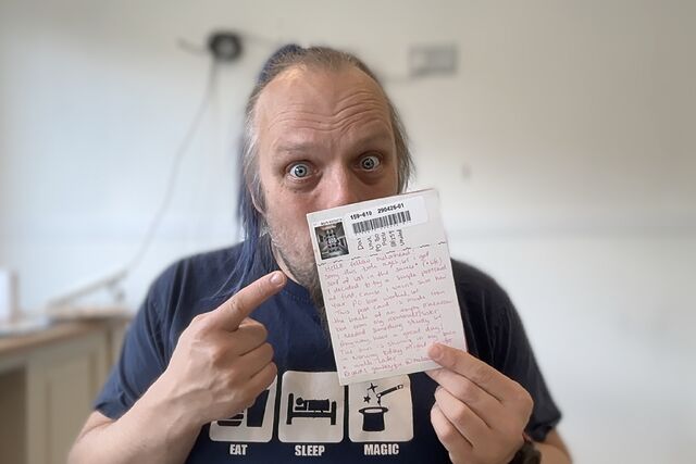 Dan, a white pan with a blue ponytail, holds a postcard in front of his face.