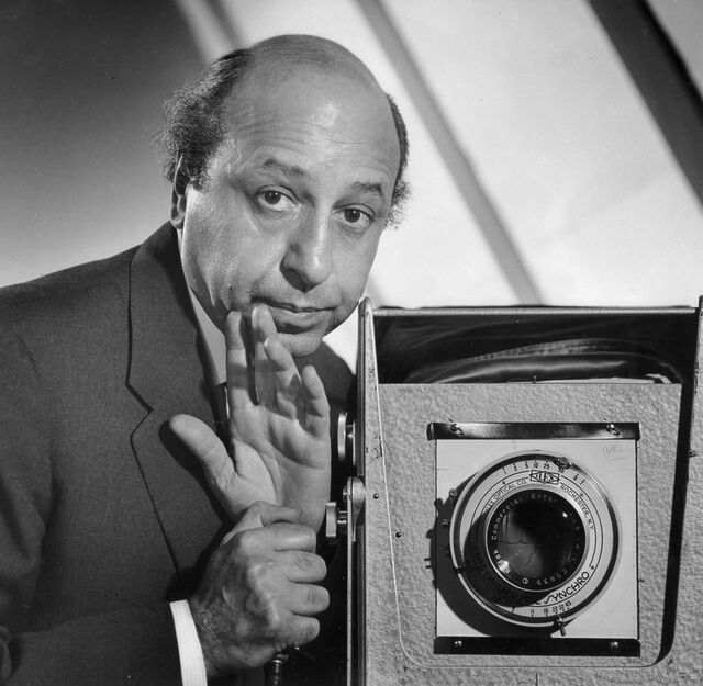 Monochome photo, presumably taken using a mirror, of a balding man operating a box camera.